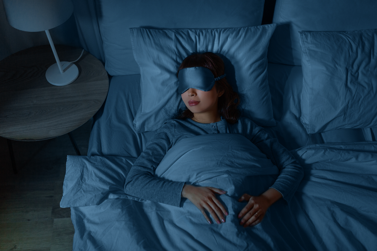 Person sleeping peacefully in bed wearing an eye mask, surrounded by blue bedding in a dimly lit bedroom at night.