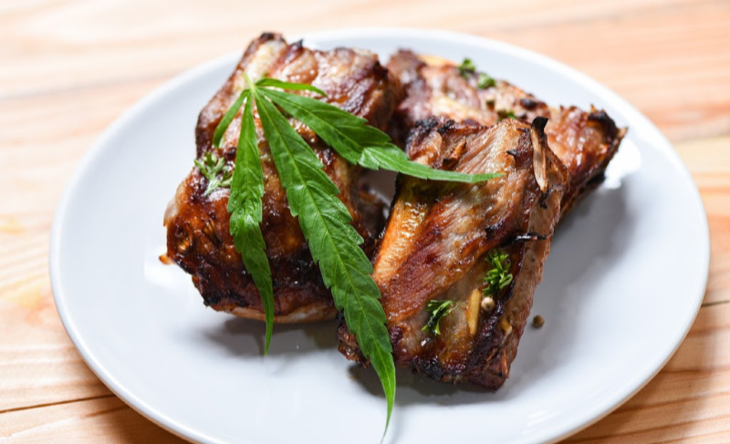 Plate of roasted meat garnished with fresh herbs and a cannabis leaf, presented on a white dish over a wooden table.