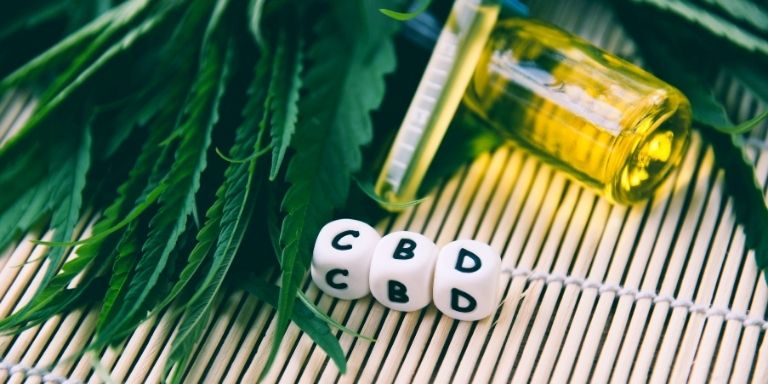 CBD oil bottle and capsules labeled CBD placed on a bamboo mat with green cannabis leaves.