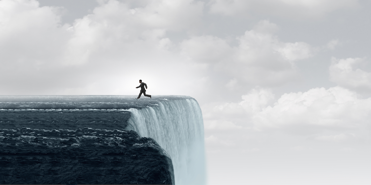 Businessman running along the edge of a massive waterfall cliff under a cloudy sky, symbolizing risk, danger, or a critical turning point.