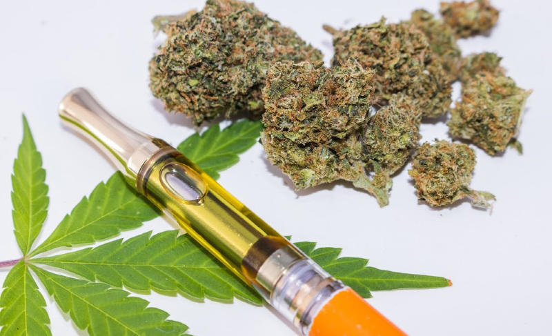 Cannabis vape pen cartridge with golden oil placed on a green marijuana leaf beside dried cannabis buds on a white background.