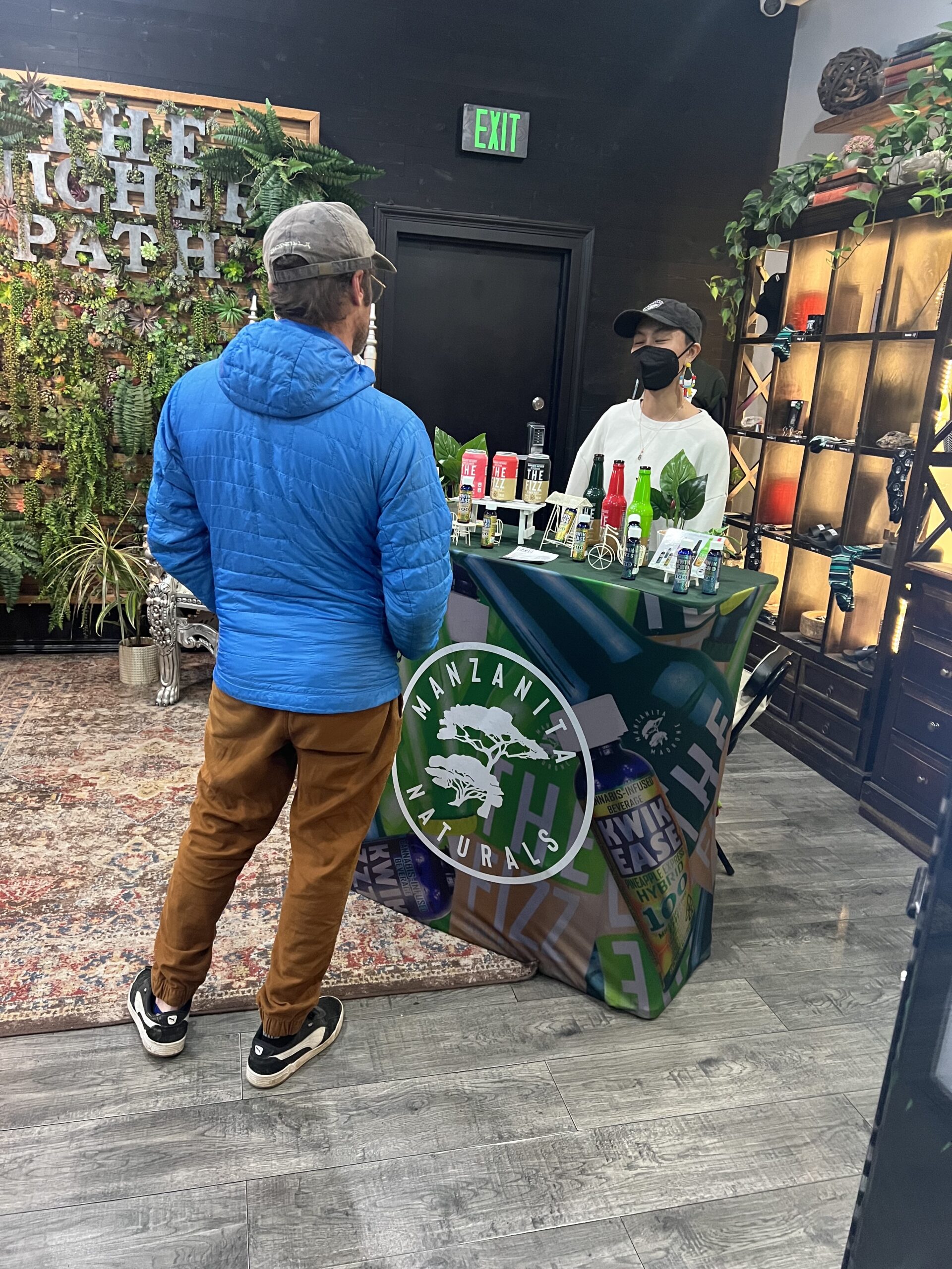 Customer speaking with a vendor at a Manzanita Naturals display table inside a plant-filled retail space, showcasing bottled beverages and wellness products.