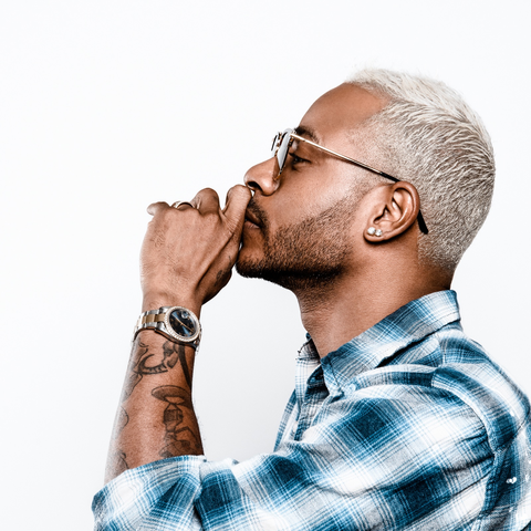 Side profile of a man with short platinum hair and glasses holding his hand near his mouth against a white background.