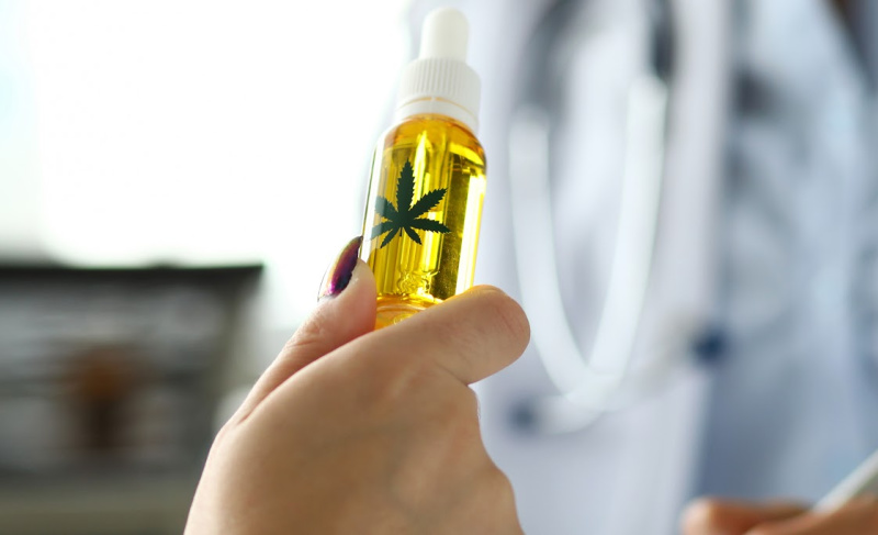Hand holding a dropper bottle filled with golden cannabis oil, featuring a cannabis leaf symbol on the label, with a medical professional blurred in the background.