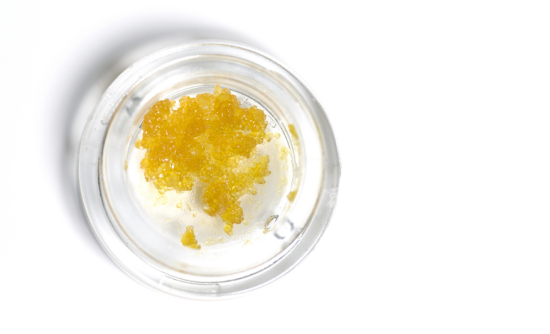 Top-down view of golden cannabis concentrate crystals inside a small clear glass container on a white background.