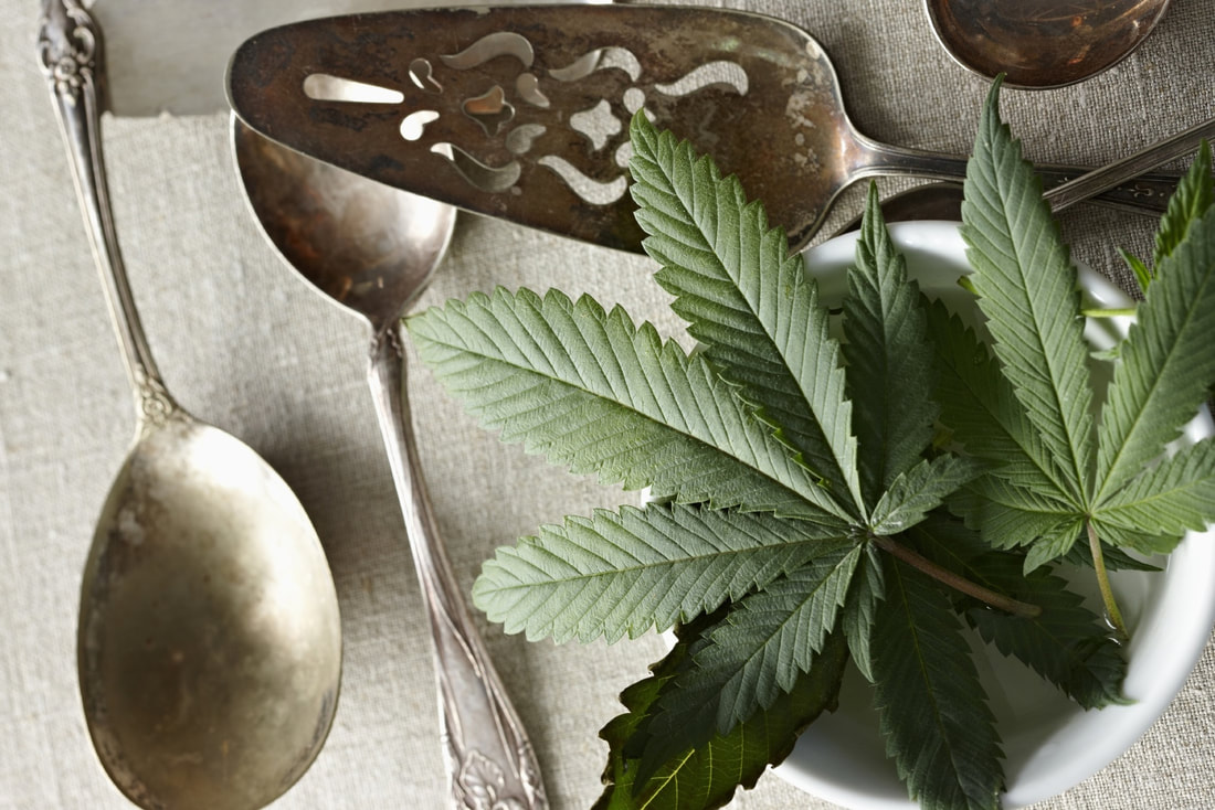 Fresh green cannabis leaves arranged in a white bowl beside vintage silverware on a neutral linen surface, suggesting cannabis use in cooking or food preparation.