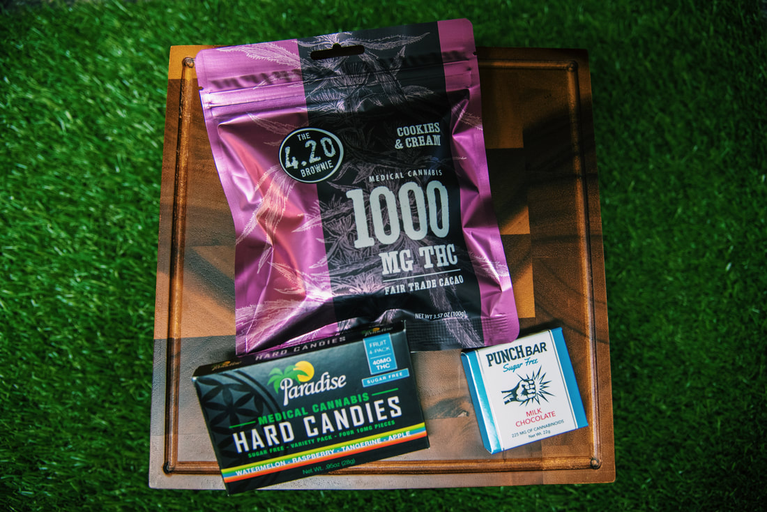 Overhead view of packaged cannabis edibles, including a brownie, hard candies, and a chocolate bar, arranged on a wooden board against a green grass background.