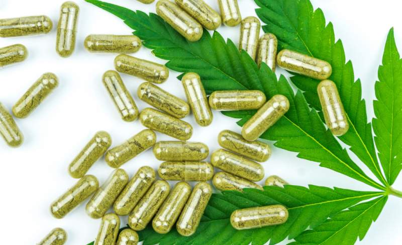 Cannabis capsules scattered on green marijuana leaves against a white background.