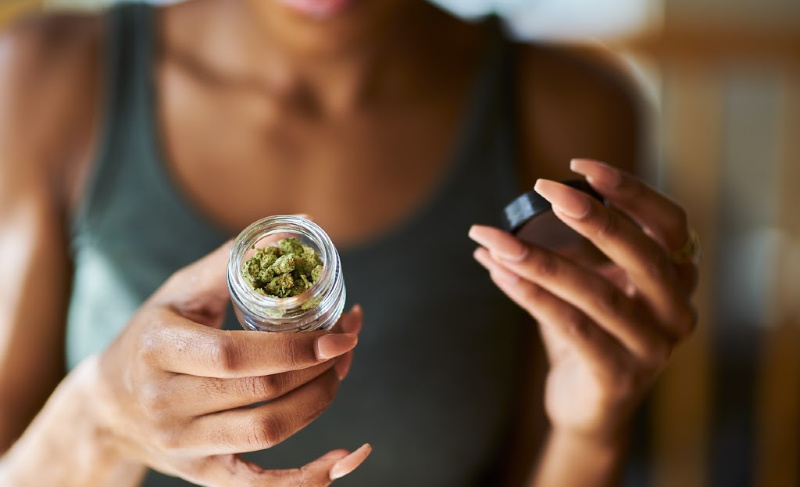 Person holding an open glass jar of cannabis flower, examining the buds with the lid in hand.