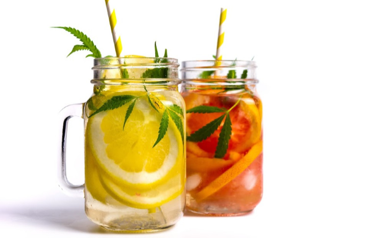 Two mason jars of cannabis-infused lemonade and fruit drink garnished with lemon slices and marijuana leaves.