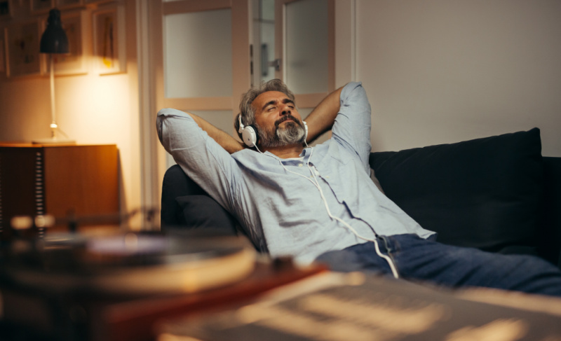 Middle-aged man relaxing on a sofa at home with eyes closed, wearing headphones and listening to music in a calm, comfortable living room setting.