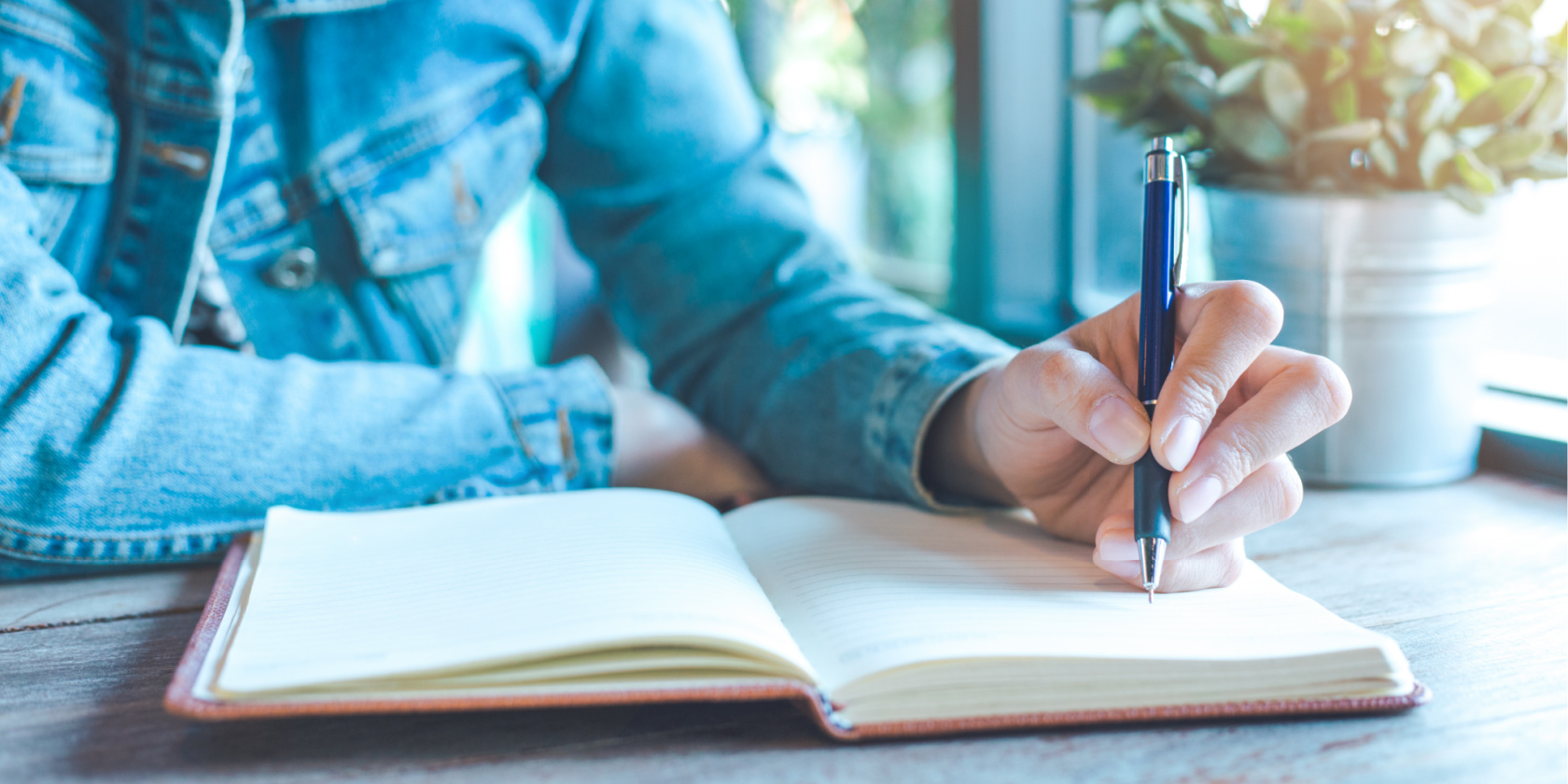 Close-up of a person writing in an open notebook with a pen at a desk near a window, suggesting journaling, note-taking, or reflective writing.