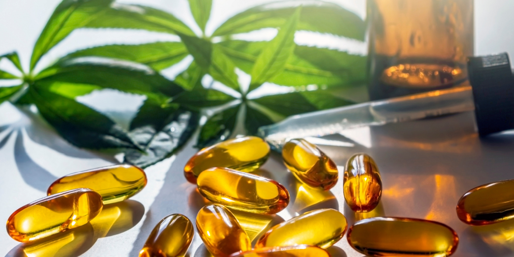 Cannabis oil capsules scattered beside a dropper bottle and green cannabis leaves on a bright surface.
