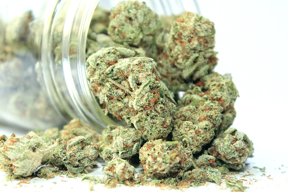 Close-up of cannabis buds spilling from a clear glass jar onto a white surface.
