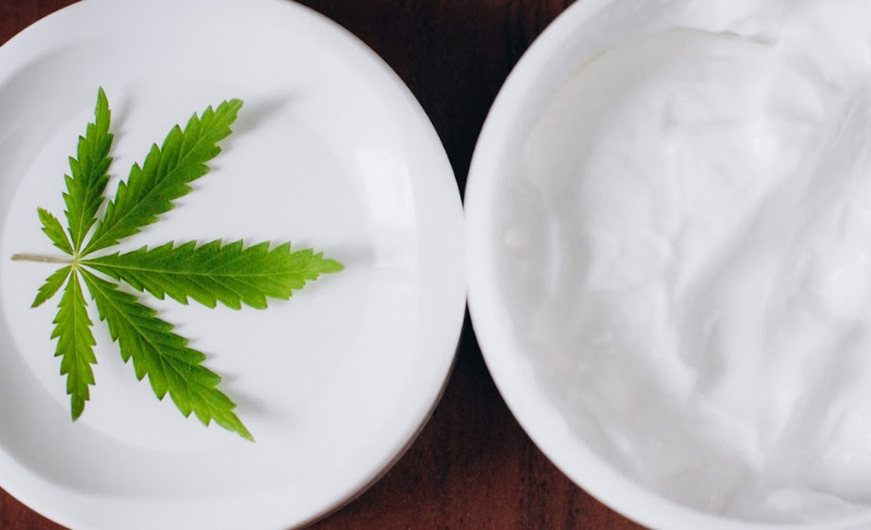 Cannabis leaf on a white plate beside a bowl of white topical cream on a dark surface.