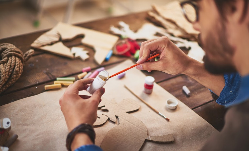 Person painting a wooden craft project at a worktable with paint, brushes, and cut-out shapes.