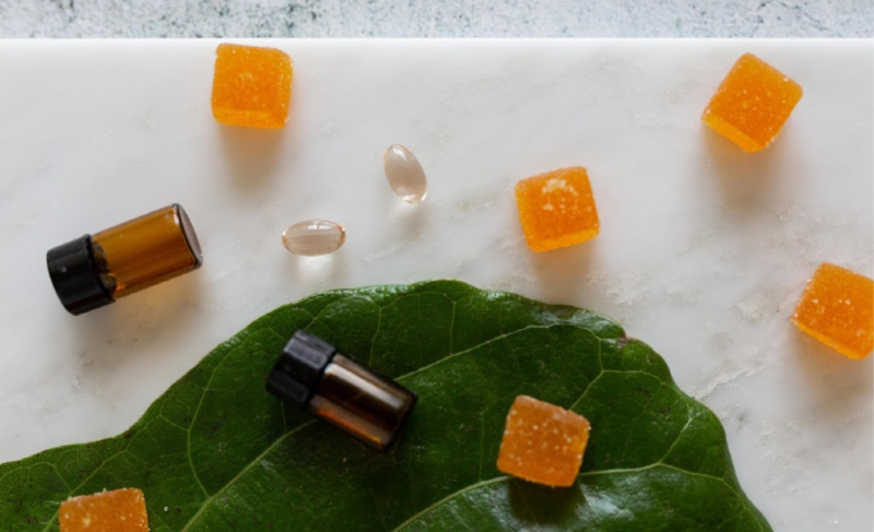 Cannabis wellness items arranged on a marble surface, including orange gummy cubes, softgel capsules, small amber bottles, and a green leaf.