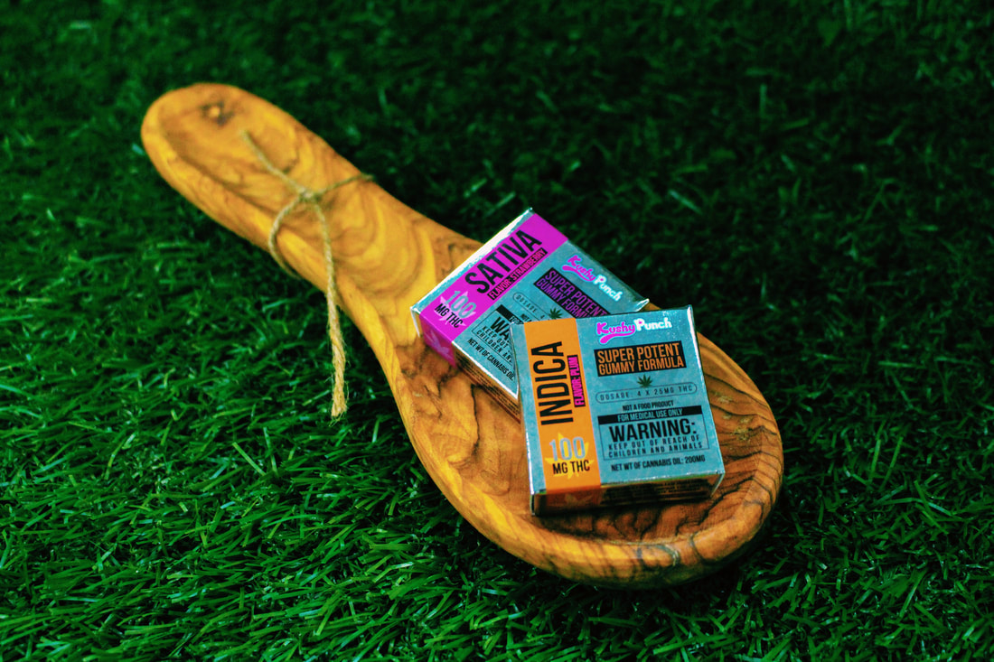 Cannabis gummy packages labeled sativa and indica resting on a wooden spoon placed on green grass.