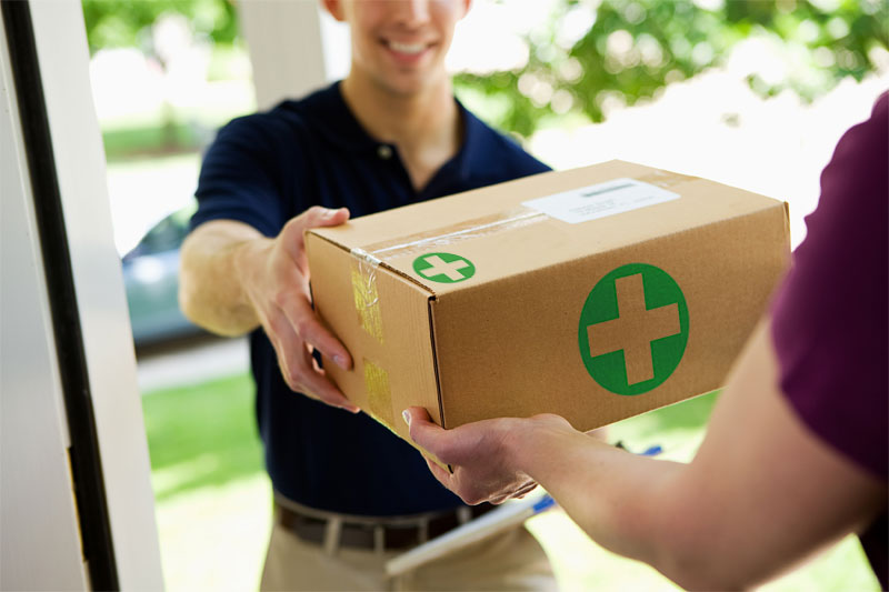 A close-up shot of a delivery person handing a cardboard box to a customer at a doorway. The box is marked with green medical cross symbols, suggesting a delivery of medical supplies or prescriptions.