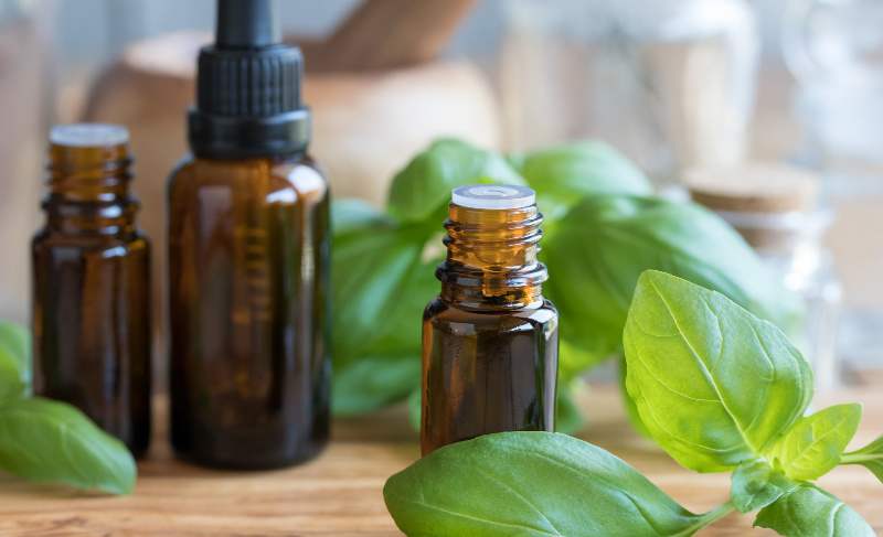 Three brown glass dropper bottles of various sizes placed among fresh green basil leaves on a wooden surface.