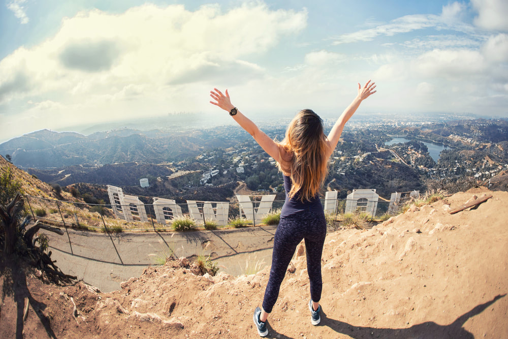 A woman stands on a hilltop with her arms raised, looking out over the Hollywood sign and the city of Los Angeles.