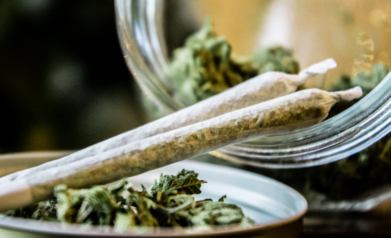 A close-up of two hand-rolled cannabis joints resting on the edge of a glass jar containing dried flower buds.