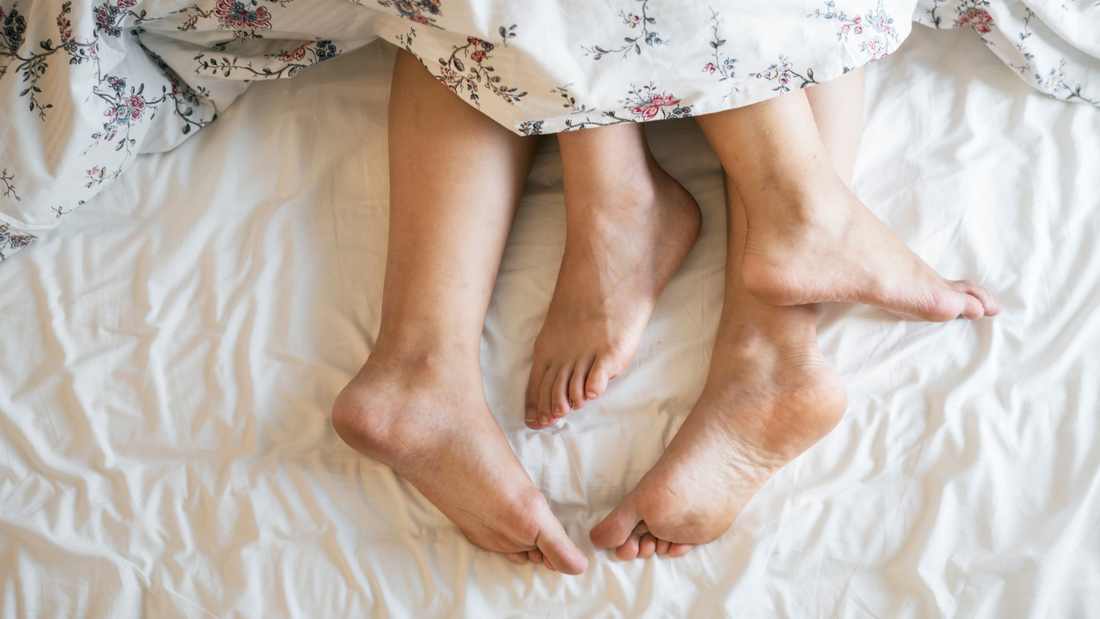 Two pairs of bare feet peek out from under a floral-patterned duvet on a rumpled white bedsheet.