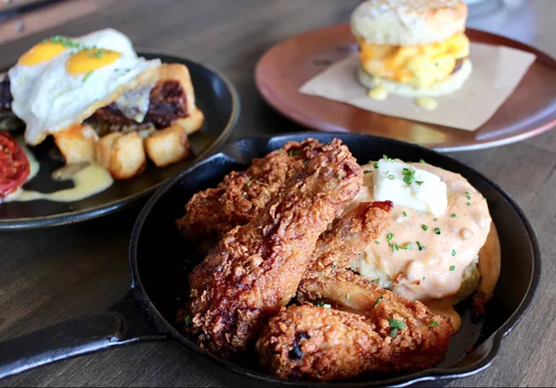 A cast iron skillet filled with crispy fried chicken legs next to a biscuit topped with gravy and butter, with other breakfast dishes visible in the background.