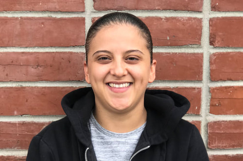 Smiling person wearing a black hoodie standing in front of a red brick wall.