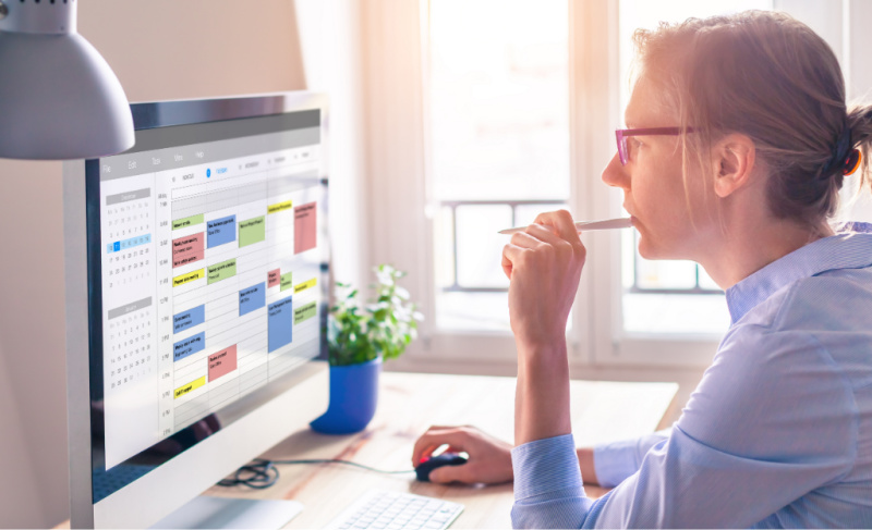 Person wearing glasses works at a desk, reviewing a color-coded calendar on a computer screen while holding a pen thoughtfully.