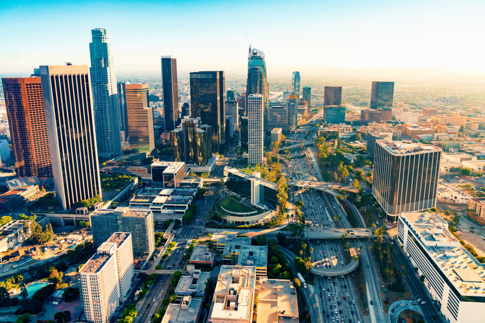 Aerial view of downtown Los Angeles skyline with high-rise buildings, busy freeways, and city streets under bright daylight.