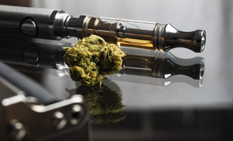 Vape pen with a cannabis oil cartridge lying next to a cannabis bud on a glossy, reflective surface.