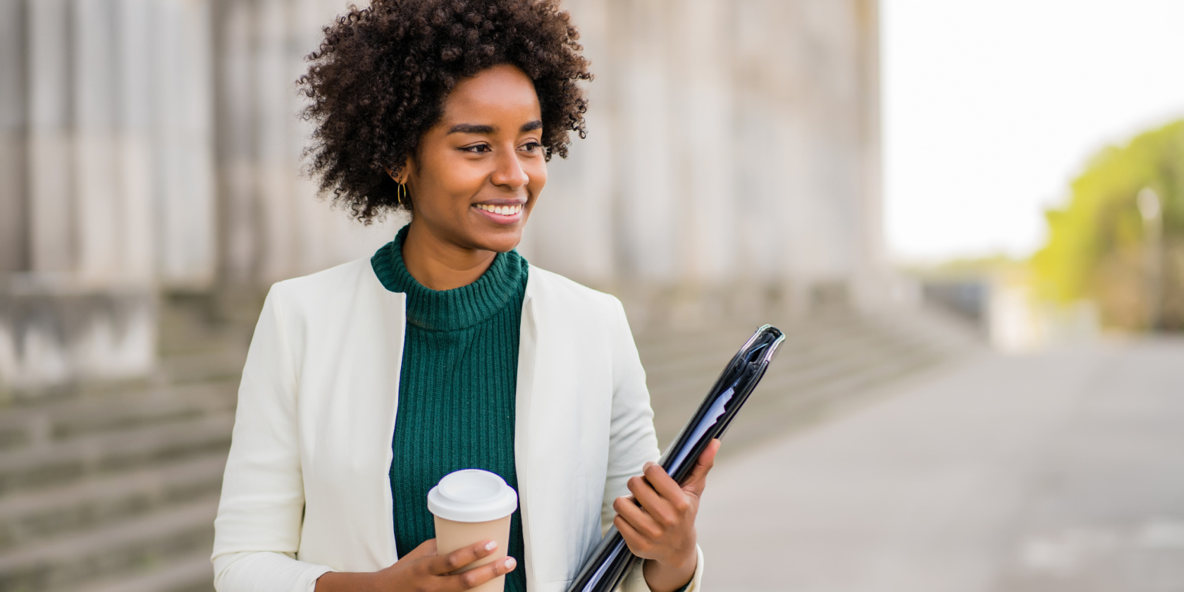 Smiling woman holding a takeaway coffee cup and a folder, standing outdoors near stone steps and columns.