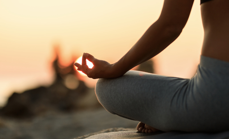 Person sitting cross-legged outdoors at sunset, hand resting on knee in a meditation mudra.